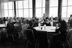 Black and white photo of a group of people seated at round tables in a large room with floor-to-ceiling windows, listening to a speaker at the front. Several attendees have their hands raised.