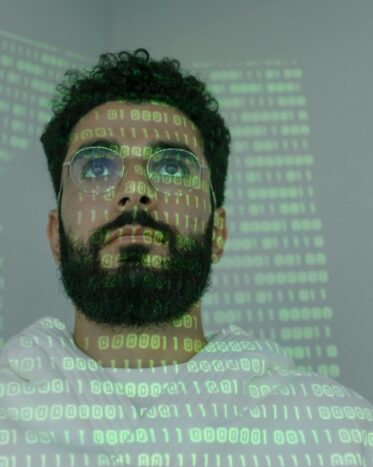 Man with a beard and glasses with binary code projected on his face and background, symbolic of data analytics